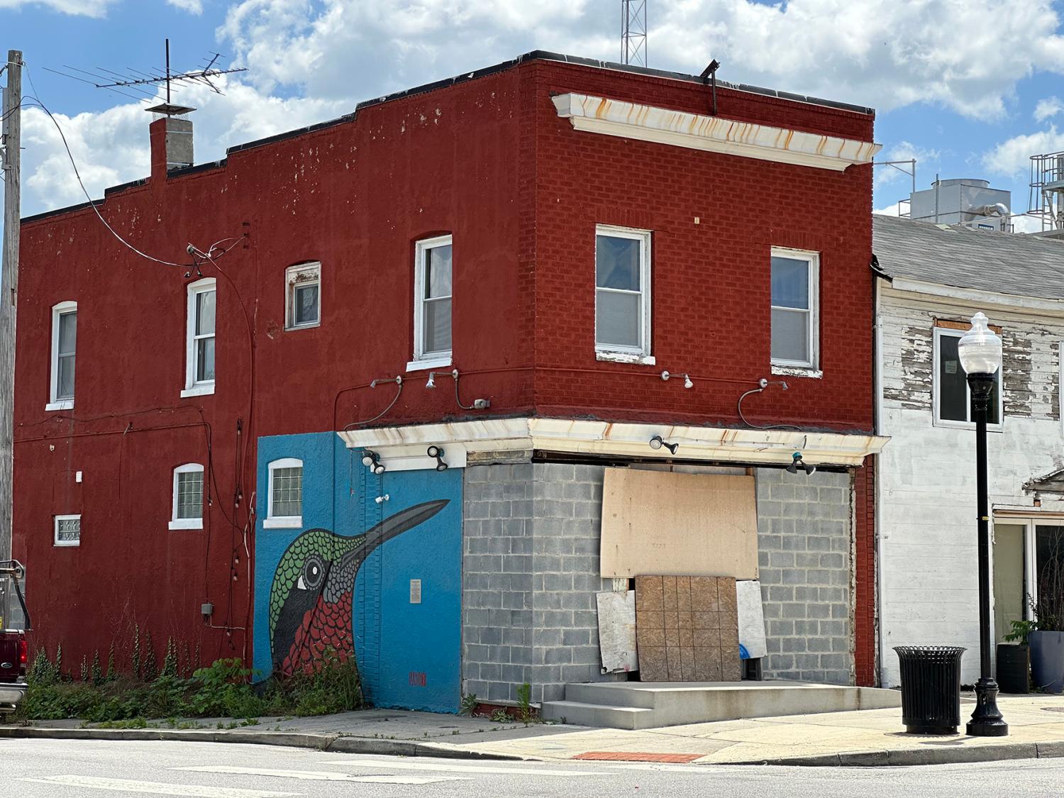 Red brick building on corner of street with hummingbird mural on exterior side wall, boarded up door