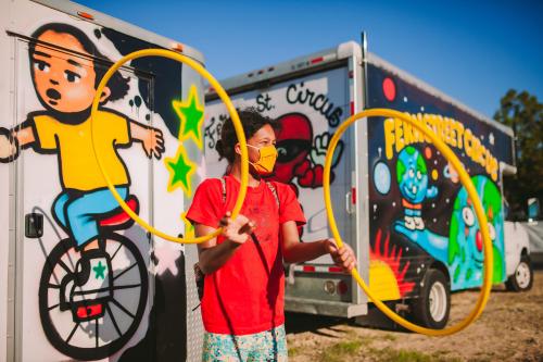 Young girl playing with two yellow hula hoops in front of brightly painted trucks advertising the Fern Street circus